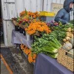 A man stands in front of a table filled with fresh vegetables, including a variety of oranges and broccoli.