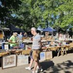 A man walks past a table full of items, including a painting, as he enjoys a day outdoors.