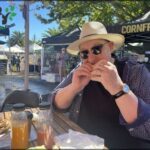 A man wearing a hat and sunglasses is enjoying a sandwich at an outdoor event.