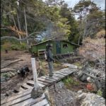 A man with a backpack walks across a bridge in front of a cabin.