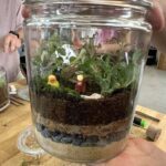 A person holds a glass container filled with a plant and a duck.