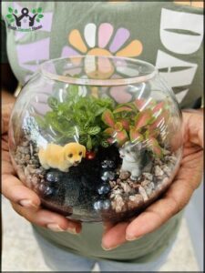 A person is holding a glass bowl with a plant and two figurines inside.