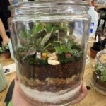 A person is holding a glass jar filled with a variety of plants and a bird figurine.