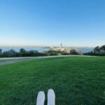 A person is sitting on a grassy field, wearing white shoes, and looking out over a city.