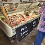 A person is standing in front of a meat display, which includes pork bacon.