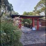 A red and white archway is surrounded by a variety of plants, including a large bush and a tree.