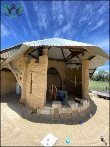 A rustic stone building with a tin roof and a chair and table inside.