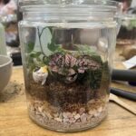 A small glass vase filled with dirt and a variety of plants, including a cat figurine, sits on a table.