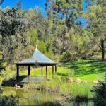 A small pavilion sits in the middle of a grassy field, surrounded by trees and a small pond.