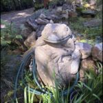 A statue of a frog is sitting on a rock in a garden.