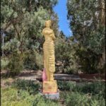 A statue of a woman is located in a park, surrounded by trees and bushes.