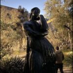 A statue of a woman with her hand on her chin is located in a park.