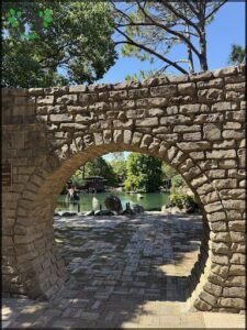A stone archway leads to a serene lake, where a group of people are enjoying the view.
