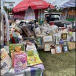 A table full of books and other items is set up under a red umbrella.