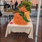 A table full of carrots and other vegetables is set up outside, possibly for a farmers market.