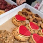 A table full of cookies, including heart-shaped ones, and a person reaching for one.