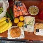 A table full of various fruits and vegetables, including bananas, oranges, carrots, and olives.