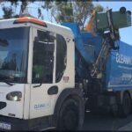 A white truck with a blue bin on the back is parked on the side of the road.