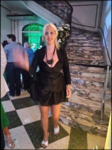 A woman in a black dress is standing on a checkered floor, waving at the camera.