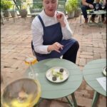 A woman in a blue apron is sitting at a table with a plate of food and a wine glass.
