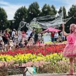 A woman in a pink dress blows bubbles in a garden filled with flowers.