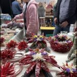 A woman in a pink jacket stands in front of a table filled with flowers and red peppers.