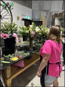 A woman in a pink shirt is standing in front of a table filled with flowers.