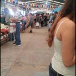 A woman in a white tank top is shopping at a market, surrounded by other shoppers and various fruits and vegetables.