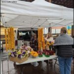 A woman is standing in front of a table filled with flowers, including yellow and orange ones. She is wearing a white jacket and appears to be selling the flowers.