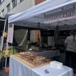 A woman is standing in front of a table filled with various pastries, including donuts and cakes.