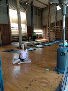 A woman sits on the floor in a room with a potted plant and several mats.
