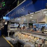 A woman stands behind a deli counter, selling cheese and other food items.