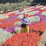 A woman stands in a garden with a heart-shaped sticker on her face, surrounded by a vibrant display of flowers.