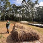 A woman stands next to a large pile of hay, possibly in a field or a barn.