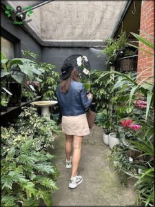 A woman wearing a hat and carrying a handbag walks through a garden filled with various plants.