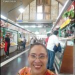 A woman wearing glasses and a colorful shirt is smiling in a marketplace.