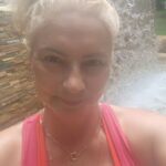 A woman with blonde hair and a pink shirt is standing in front of a waterfall, smiling for the camera.
