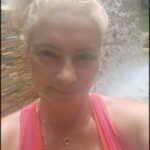 A woman with blonde hair and a pink tank top poses for a picture in front of a waterfall.