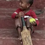 A young boy is sitting on the stairs, holding a tennis ball and a baseball bat.