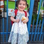 A young girl wearing a blue and white dress is holding a hot dog in her hand.