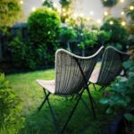 Two chairs sit in a garden, surrounded by greenery and lights.