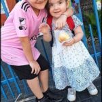 Two children pose for a picture, one holding a pastry and the other holding a backpack.