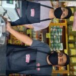 Two men wearing face masks are standing in front of a deli counter, likely preparing food.