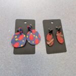 Two pairs of earrings are displayed on a table, one with a blue and yellow design and the other with a red and black design.