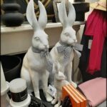 Two white bunny statues sit on a table with a book and a hat.