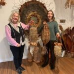 Two women are standing in front of a wall display, each holding a woven object.