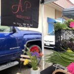 A blue truck is parked in front of a flower stand, with a sign advertising the sale of flowers.