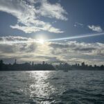 A boat is sailing on the water in front of a city skyline, with the sun shining brightly in the sky.