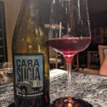 A bottle of Cara Siliga sits next to a glass of red wine on a table.