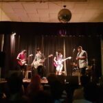 A group of four people are playing instruments on stage, with a crowd of people watching them.